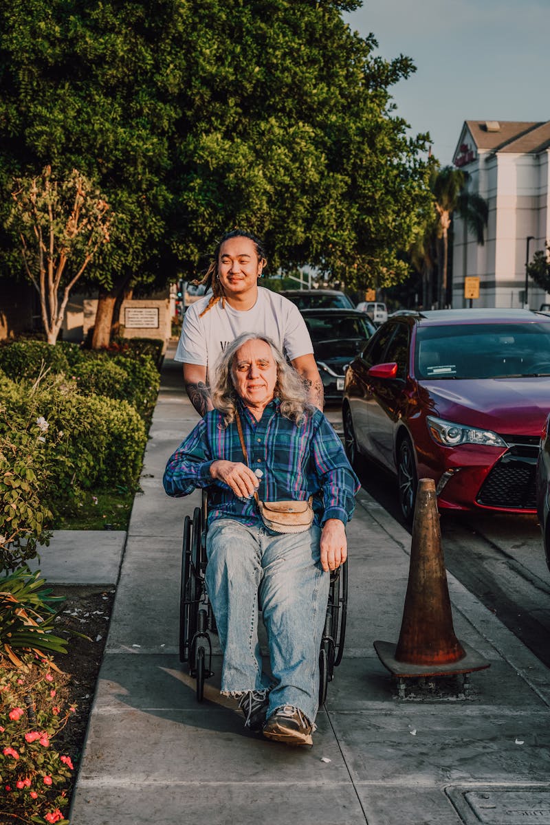 A caring scene of a young man pushing a senior in a wheelchair along a sidewalk.